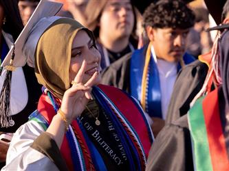 A female graduate wearing a gold hijab and a "Seal of Biliteracy" stole smiles and gives a peace sign to the crowd.