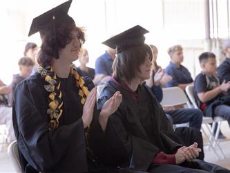 Two graduates in caps and gowns sit next to each other and applaud during a presentation, with one wearing a black-and-gold money lei. Other audience members sit in rows behind them.