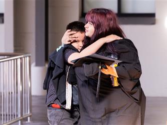 A graduate with long, purple-dyed hair wearing a graduation gown embraces a young boy in a heartwarming hug. She is holding her graduation cap and a clear cup filled with colorful items in her hand.