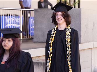 A smiling graduate with wavy brown hair wears a black cap, gown, and an elaborate gold-and-black ribbon lei woven with folded dollar bills. He walks in a procession behind a graduate with reddish-purple hair.