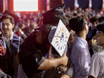 Two graduates share a warm embrace in a crowded stadium at night, with one wearing a cap decorated with "Foothill Family."