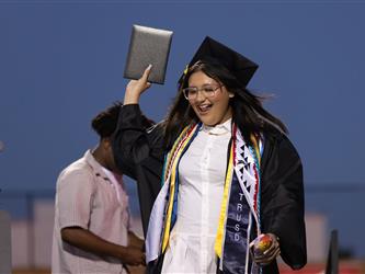 A female graduate wearing glasses and a black gown smiles excitedly while raising her diploma cover high in the air on stage.
