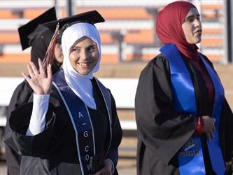 A smiling graduate wearing a white hijab and black gown waves her hand to the side while walking outdoors during the graduation ceremony.