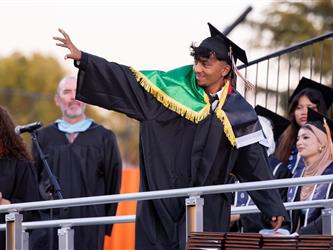 A male graduate wearing a black gown with a green, gold-fringed shoulder overlay smiles and waves to the crowd from a stage.