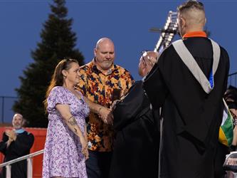 A man in a floral shirt and a woman in a purple dress shake hands with school officials on stage during a ceremony.