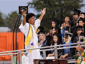 A graduate in a white gown and yellow stole stands on stage with arms raised, cheering and holding up his diploma cover in front of seated classmates.
