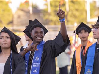 A smiling male graduate in a black gown and blue stole points one finger high in the air while standing with classmates outdoors.