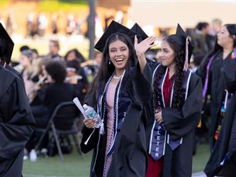 A female graduate with long dark hair smiles excitedly and waves to the crowd while walking in a procession on a sports field.