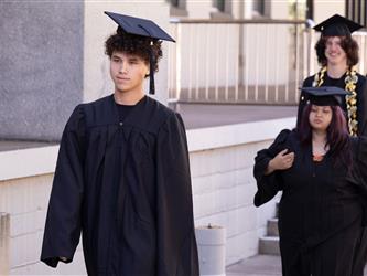 A graduate with curly dark hair wearing a black cap and gown walks forward during a processional ceremony. Other graduates in matching caps and gowns follow close behind him along an outdoor concrete walkway.