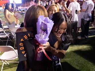 A female graduate smiles warmly while hugging a friend and holding a purple bouquet at an outdoor graduation venue at night.