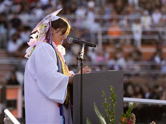 A female graduate in a white gown stands at a podium reading her speech to a crowd seated in stadium bleachers.