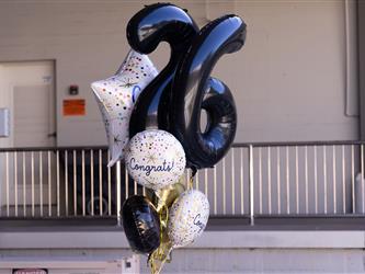 A festive bouquet of helium balloons floating outdoors near a loading dock ramp. The arrangement includes large black foil balloons shaped like the numbers "2" and "6," a star-shaped balloon with colorful confetti dots, and several round white balloons printed with colorful confetti and the word "Congrats!