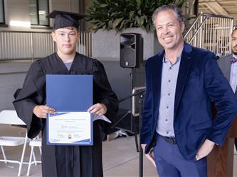 A young male graduate wearing a black cap and gown holds open a blue folder displaying a certificate. To his right, a man with curly graying hair in a blue velvet jacket smiles broadly. A man in a gray suit and bowtie stands behind a wooden podium in the background.