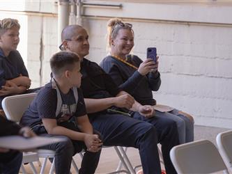 Graduates family members sitting in rows of white folding chairs at an outdoor ceremony. In the center, a woman smiles while holding up her purple smartphone to take a photo, seated next to a man and a young boy who are watching the event.