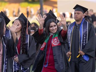 A smiling female graduate wearing an Afghan flag-themed stole waves both hands in the air during a graduation procession.