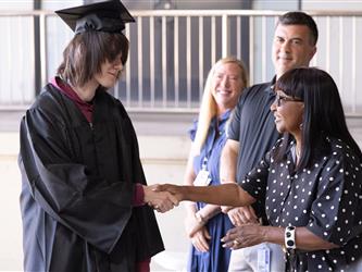 A side-profile view of a graduate with long dark hair and a graduation cap and gown shaking hands with a smiling woman wearing glasses and a black dress with white polka dots. Two other onlookers, a man and a woman, stand in the background smiling.
