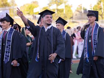 A male graduate in a black cap and gown points forward with a smile while walking with classmates outdoors
