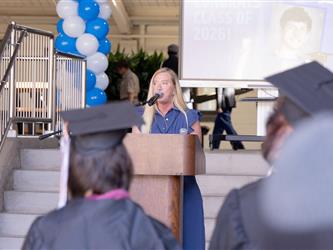 A blonde woman in a ruffled blue dress speaks into a microphone at a wooden podium on a stage. The view is framed from behind two seated graduates wearing caps and gowns, with a blue and white balloon column visible to the left.