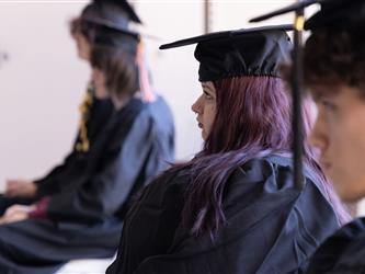 A profile view of three graduates sitting side-by-side in a row during their commencement ceremony, all wearing black graduation caps and gowns.