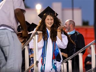 A female graduate in a white gown and colorful honor cords smiles brightly and waves both hands toward the crowd while walking across the stag