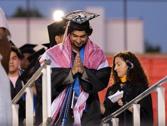 A graduate wearing a black gown and a red-and-white patterned Keffiyeh walks down the stage steps with hands pressed together in a respectful gesture.