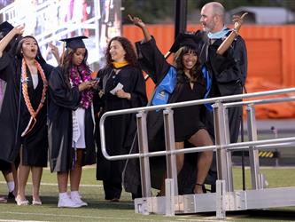 A smiling female graduate walks down the stage ramp dancing with her arms up, while school administrators and cheering fellow graduates look on.