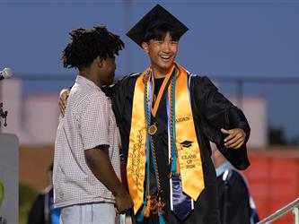 A smiling male graduate wearing an orange "Class of 2026" stole stands on stage with his arm around a friend in a plaid shirt.