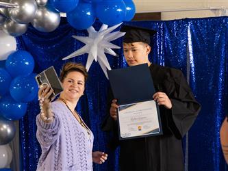 A woman in a lilac knit cardigan holds up her phone to take a selfie with a young male graduate in a cap and gown. The graduate is holding open his blue certificate folder, standing in front of a blue and silver balloon backdrop.