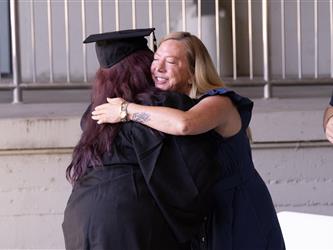 A blonde woman in a dark blue dress shares a warm hug with a graduate wearing a black cap and gown.