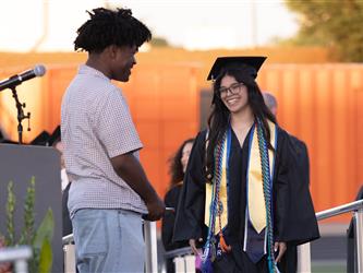 A smiling female graduate with glasses and a yellow stole walks across a stage toward a school staff member
