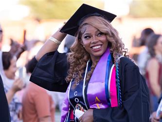 A smiling female graduate with curly blonde hair looks to the side while adjusting her black mortarboard cap in a crowd.