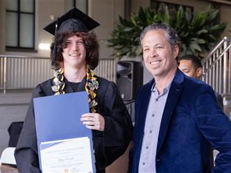 A young graduate with long, dark, wavy hair wearing a black graduation gown, cap, and a gold and black money lei. He is smiling and holding up a blue folder containing a certificate that reads "Certificate of Participation presented to Riley Privett." Standing next to him on the right is a smiling man with graying hair wearing a blue velvet blazer over a gray button-up shirt.