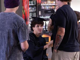 A young graduate with curly dark hair wearing a graduation cap and gown sits and shakes hands with a friend. He is surrounded by a small group of friends talking in an indoor hallway setting.