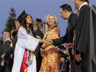A female graduate in a white gown smiles while shaking hands with a school official on stage during a graduation ceremony.