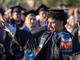 A male graduate with curly hair waves to the side while wearing a colorful Mexican serape-style stole over his black gown.