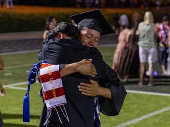 An emotional graduate in a black cap and gown closes his eyes while sharing a tight, heartfelt hug with a loved one on the field.