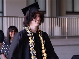 A young graduate with long, dark, wavy hair wearing a black graduation gown, cap, and a gold and black money lei walking and smiling.