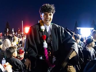 A joyful male graduate wearing a black gown and a colorful floral lei is carried on someones shoulders in a crowded celebration at night.