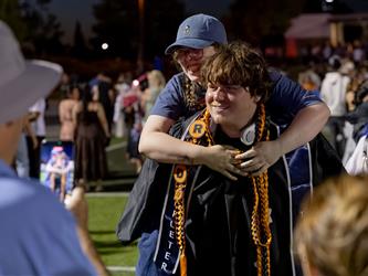 A person in a blue baseball cap gives a playful piggyback ride to a smiling graduate in a black gown while someone takes a photo.