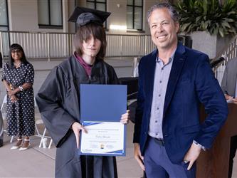 A graduate with long, straight dark hair covering part of his face stands in a black graduation cap and gown, holding open a blue folder displaying a certificate. Next to him, a man in a blue velvet blazer smiles at the camera. In the background, other ceremony attendees and a podium are visible.