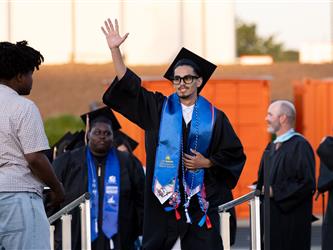 A male graduate wearing a blue stole, glasses, and a black cap waves his hand to the crowd as he walks across an outdoor stage.