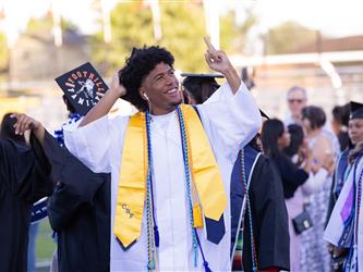 A male graduate in a white gown and yellow stole smiles and points upward with one hand while adjusting his cap with the other.