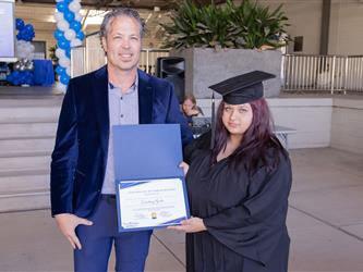 A man in a blue velvet blazer stands alongside a female graduate with purple-red hair as they jointly hold open a blue Twin Rivers Certificate of Participation folder for a photo.