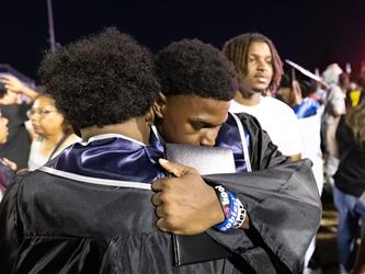 Two male graduates in black gowns share a close, emotional embrace in a crowded stadium area at night.