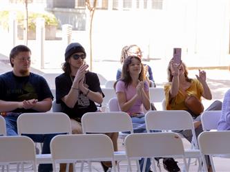 An audience seated in rows of white folding chairs outdoors. In the middle row, a young man in a beanie and a young woman in a pink shirt are clapping. Next to them, a woman in a yellow shirt holds up a smartphone to take a picture of the stage.