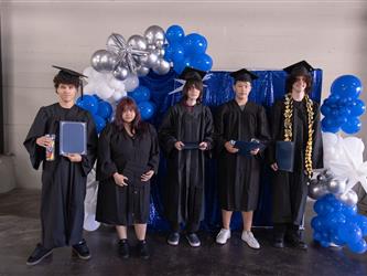 A group photo of five graduates standing in a row in front of a festive backdrop made of shiny blue fabric and an arch of blue, white, and silver balloons. They are all wearing black caps and gowns and holding their graduation folders or certificates.