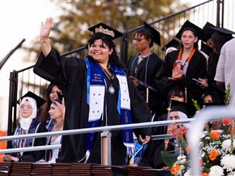 A smiling female graduate wearing a tiara on her cap and a blue and white stole waves to the crowd from an outdoor stage.