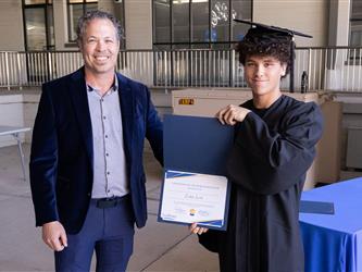 A smiling man in a blue velvet blazer and grey button-down shirt stands next to a young male graduate in a cap and gown who is holding up a blue Twin Rivers Certificate of Participation folder.
