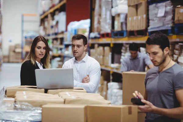 2 men and a women looking at a laptop in a warehouse full of boxes.