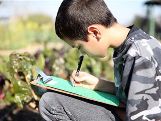 A student taking notes on a green piece of paper on a clipboard while outside in the garden